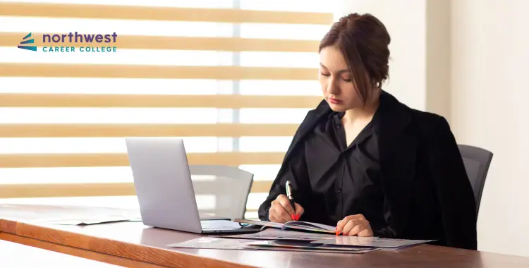 A woman in a black outfit sits at a desk, writing in a notebook with a laptop nearby and sunlight filtering through blinds.
