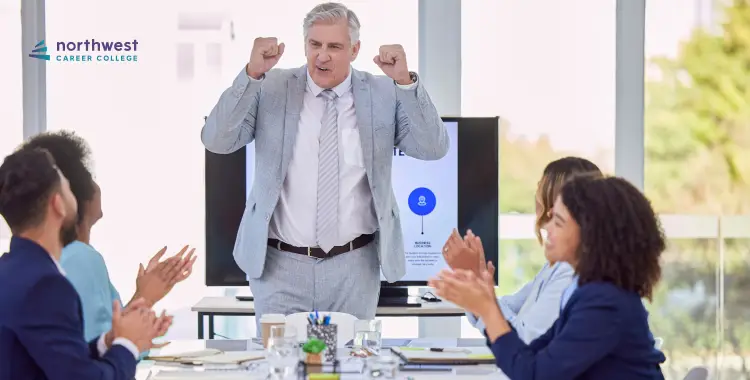 A man in a suit celebrates while colleagues applaud in a bright conference room.