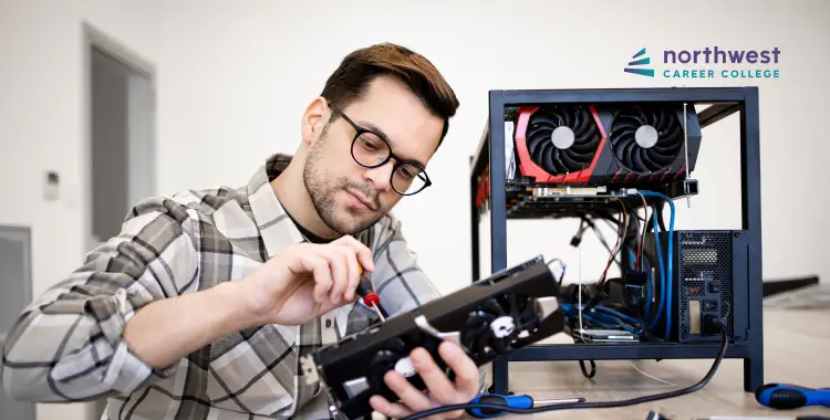 A man in glasses works on a computer graphics card, with a computer tower in the background.