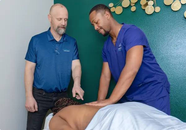 Two instructors demonstrate techniques in a massage therapy class, focusing on a student receiving a massage.