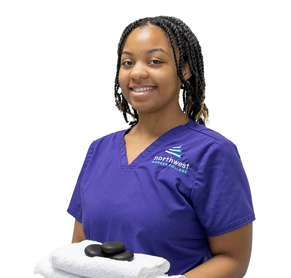 Smiling student in scrubs holds towels and stones, representing a massage therapy school environment.