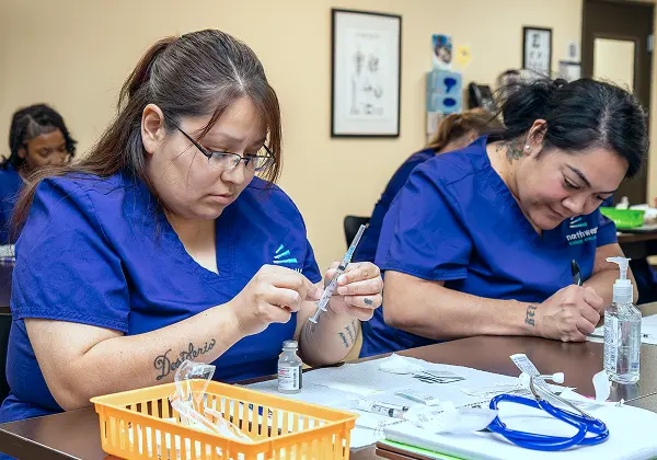 Two students in blue scrubs focus on tasks in a Medical Assistant Class, preparing syringes and practicing techniques.