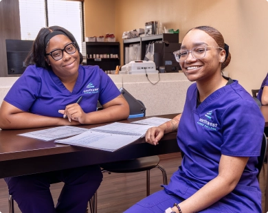 Two students in purple scrubs smile while studying together during pharmacy tech training.