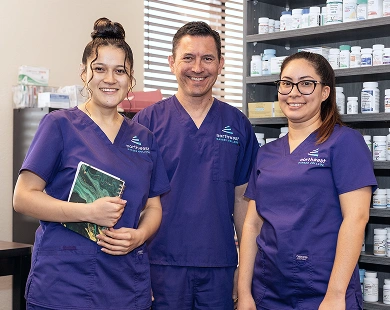 Three pharmacy tech trainees in scrubs smile together in a pharmacy setting, surrounded by shelves of medication.
