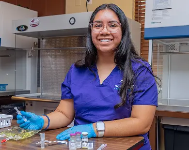 Smiling student in a lab coat engaged in pharmacy technician training, preparing vials and syringes at a workstation.