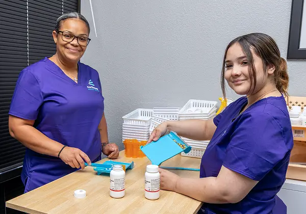 Two women in scrubs work together at a table, engaged in a pharmacy technician training program, handling medication bottles.