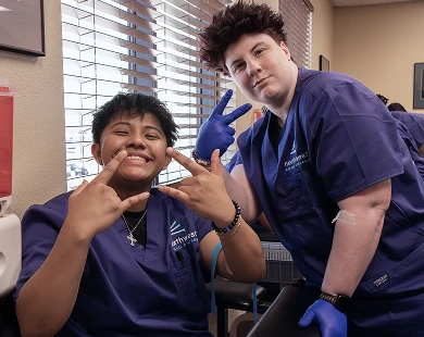 Two smiling individuals in scrubs pose playfully during phlebotomist training, showcasing a fun learning environment.
