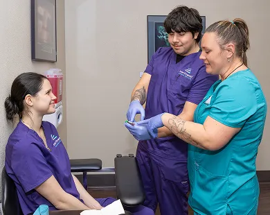 Two healthcare professionals in scrubs demonstrate phlebotomy techniques to a seated student in Las Vegas.