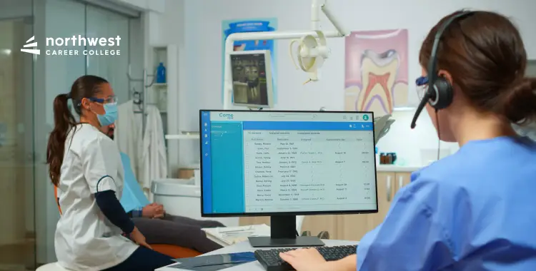 A dental office scene with a student in scrubs using a computer, while another student observes.