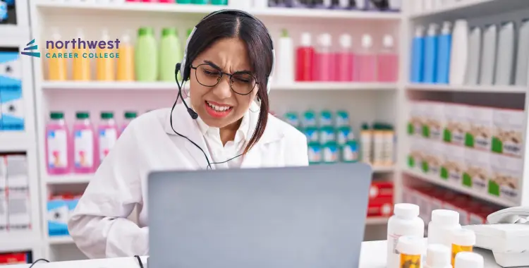A woman in a lab coat and headset looks at a laptop, surrounded by colorful bottles and medication containers.
