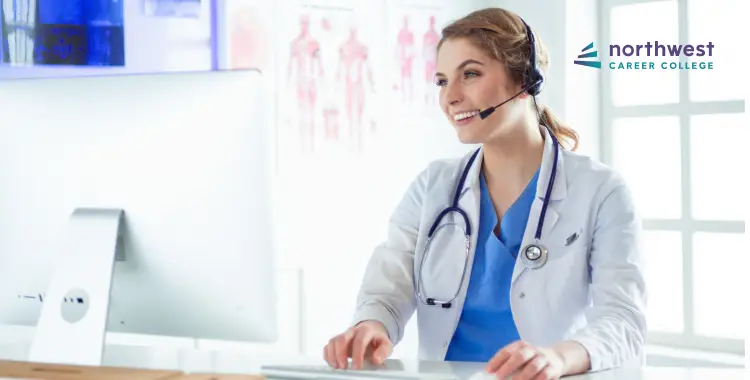 A smiling female healthcare professional in a lab coat uses a headset while working at a computer.