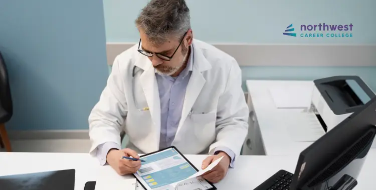 A man in a lab coat reviews documents on a tablet at a desk with a computer and printer.