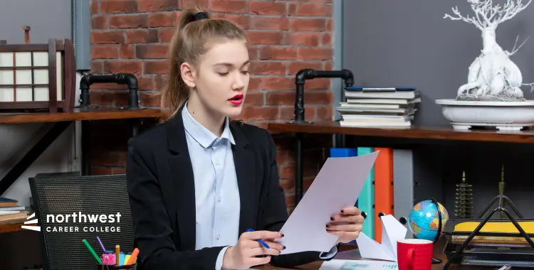 A young woman in a blazer sits at a desk, reviewing documents with colorful stationery and a globe nearby.