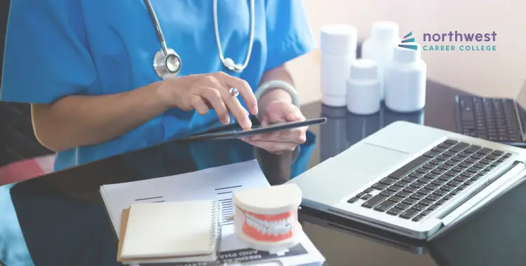 A healthcare professional in scrubs uses a tablet at a desk with a laptop, notepad, and dental model.