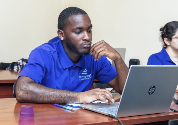Student engaged in BA Program hands-on training in class, focused on a laptop with peers in the background.