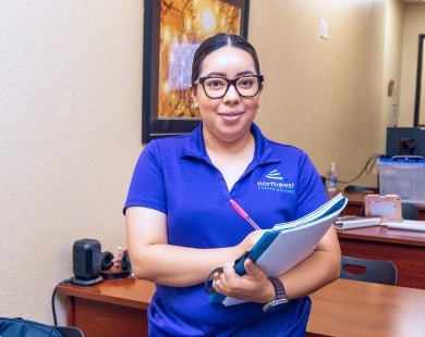 NCC BA Student holding a notebook and pen, smiling in a classroom setting with desks and supplies in the background.