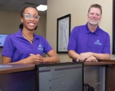 Two smiling staff members in purple shirts at a reception desk, engaging with the baa program during class.