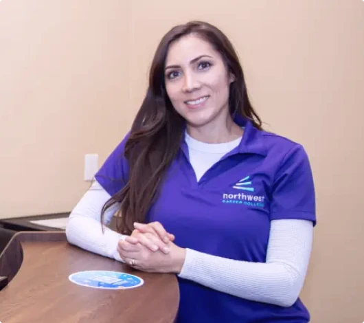 A smiling woman in a purple shirt sits at a desk, representing a Business Administration Program Student.