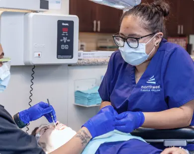 A dental student in scrubs examines a patients mouth while another person assists in a clinical setting.