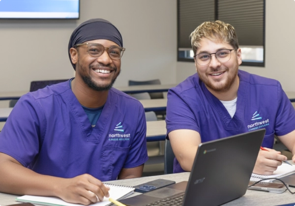 Two medical billing coding students smile while studying together at a table with a laptop and notebooks.