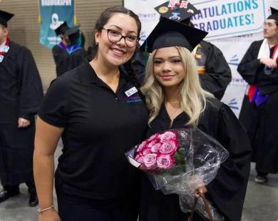 Two women smile together at a graduation event, celebrating ncc mbc graduates with flowers and caps in the background.