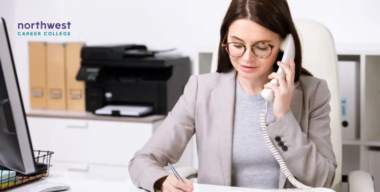 A woman in a blazer sits at a desk, talking on the phone and taking notes. Office supplies are visible in the background.