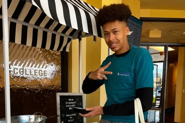 A smiling young man in a teal shirt poses by a welcome sign for new students at a college event.