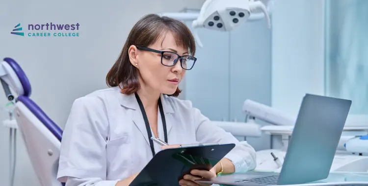 A focused woman in a lab coat reviews notes on a clipboard while working on a laptop in a dental office.