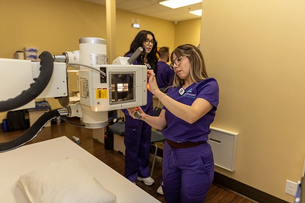 A healthcare professional operates an X-ray machine in a clinical setting, with colleagues in the background.