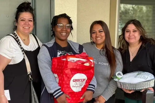 Four women stand together at a doorway, smiling and holding a red grocery bag and a tray with a turkey.