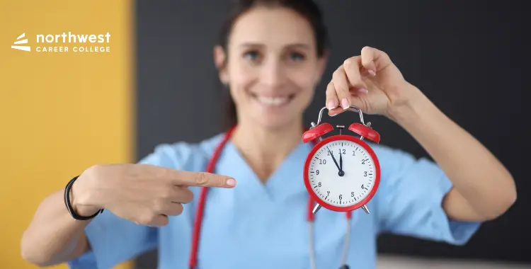 A smiling healthcare professional in scrubs holds a red alarm clock, pointing at it with enthusiasm.