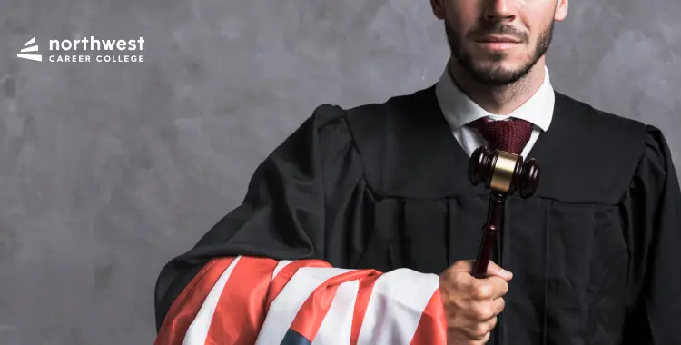 A man in a judges robe holds a gavel, draped in an American flag, against a gray background.