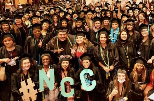 A large group of graduates in caps and gowns, celebrating with a #NCC sign in a festive indoor setting.