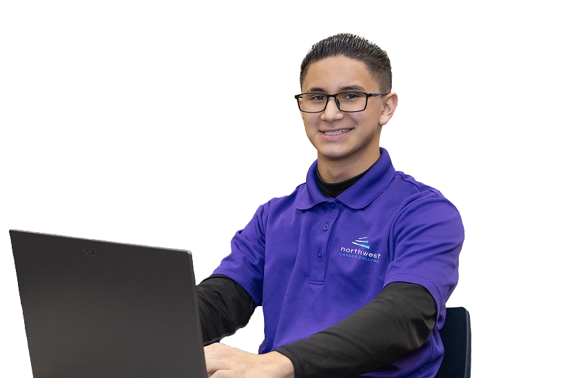 A smiling young man in a purple polo shirt sits at a laptop, looking at the camera.
