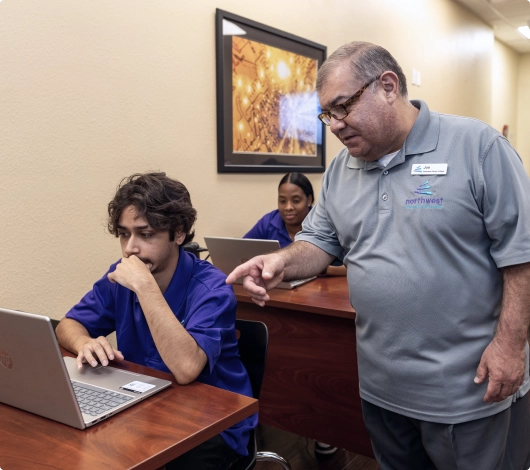 An information technology instructor assists a student at class, focusing on a laptop while another student observes.