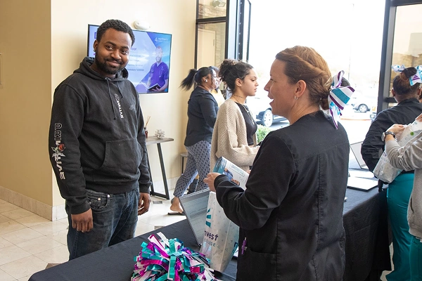 A man smiles at a table while a woman hands him a bag; others are engaged in conversation in the background.