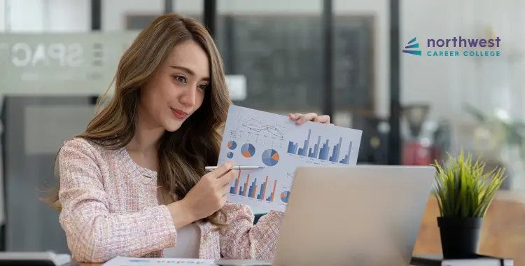 A woman analyzes graphs and charts while working on a laptop in a modern office setting.