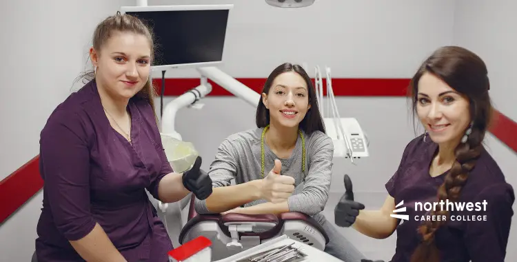 Three women in a dental office giving thumbs up, smiling, with dental tools and equipment in the background.