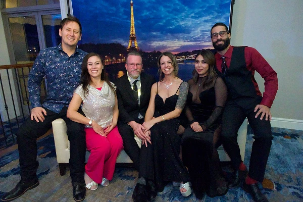 A group of six people dressed elegantly poses together in front of a backdrop featuring the Eiffel Tower at night.