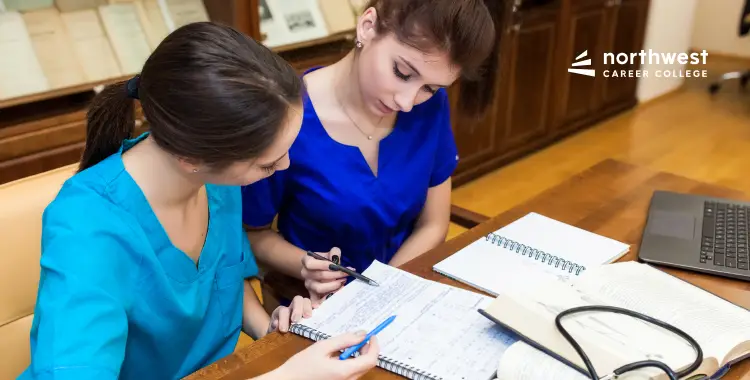 Two students in scrubs collaborate on notes at a desk, surrounded by books and a laptop.