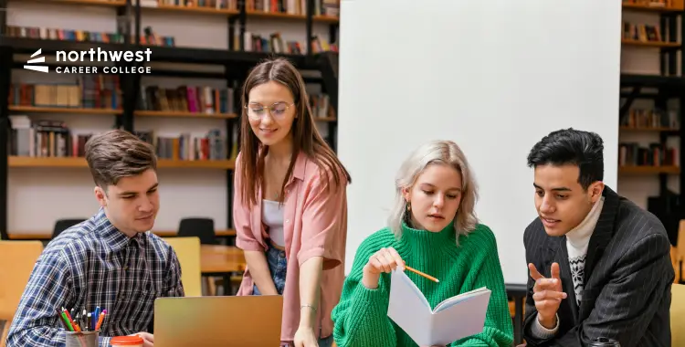 Four students collaborate around a laptop, discussing notes in a library setting.