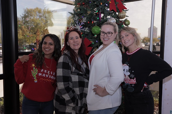Four women pose together in front of a Christmas tree, smiling and wearing festive attire.