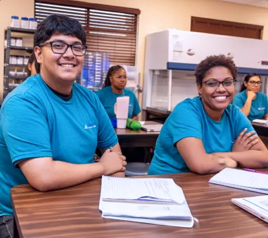 Students in a healthcare administration school classroom, smiling and engaged, with notebooks and a lab setup in the.