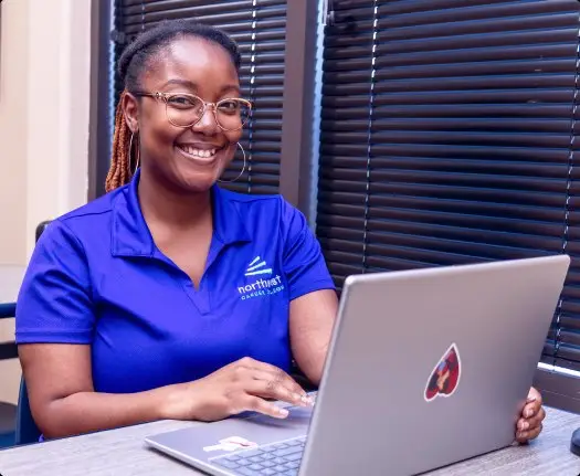 Smiling student in a blue shirt works on a laptop, pursuing a healthcare management associate degree.