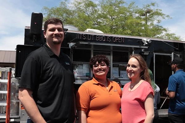 Three people stand in front of a BBQ food truck, smiling. The truck sign reads ITS LIT BBQ IS OPEN.