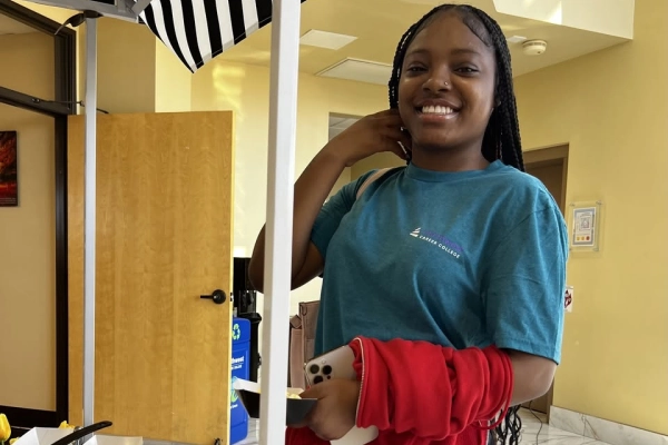 Smiling young woman in a blue shirt stands by a food station, holding a plate and phone, with a striped umbrella above.