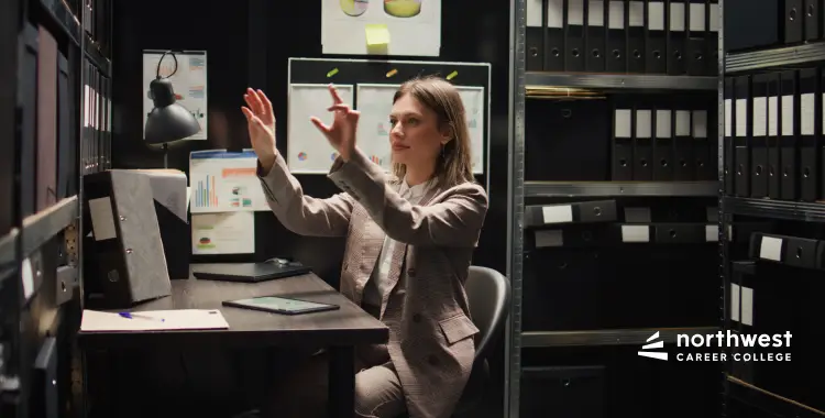 A woman in a suit gestures while sitting at a desk surrounded by filing cabinets and documents.