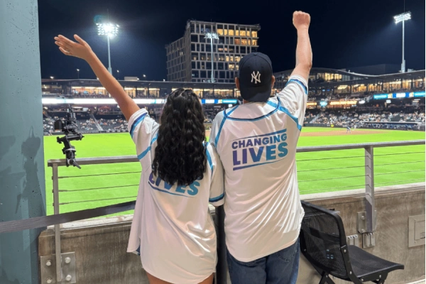 Two people in matching shirts with Changing Lives stand at a baseball game, cheering with arms raised.