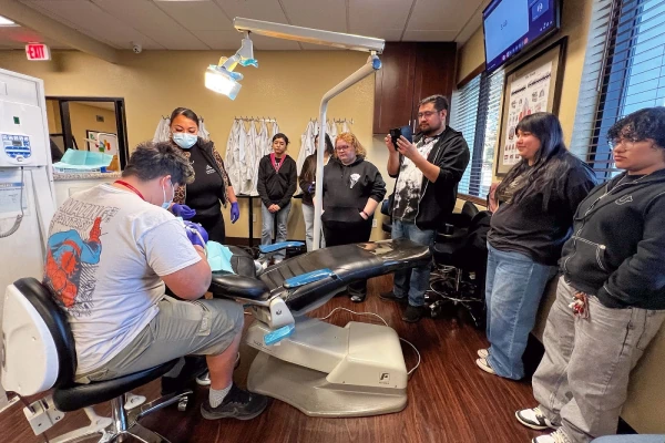 A group observes a dental procedure in a clinic, with one person in the chair and others watching attentively.
