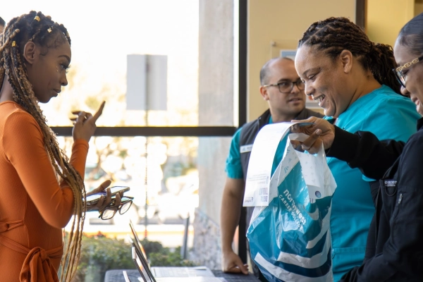 A woman in an orange dress interacts with two healthcare workers at a table, while a man observes.